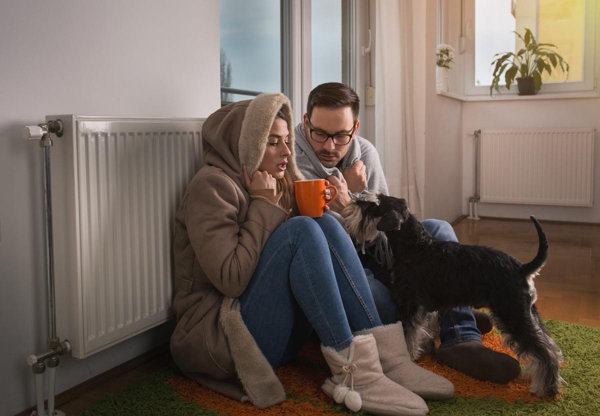 A husband and his wife trying to stay warm by leaning against the radiator in their home.